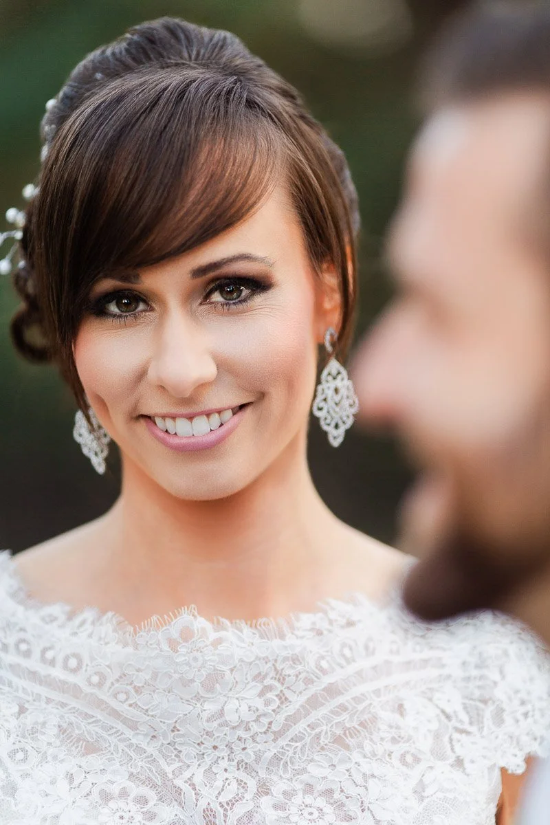 Smiling bride in lace dress and ornate earrings, with blurred groom in foreground. The mood is joyful and affectionate, set in a soft outdoor light.