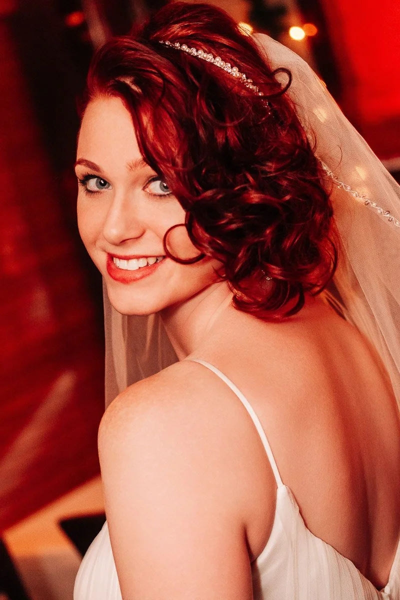 A smiling bride with red curly hair and a sparkling tiara. She's wearing a white dress and veil, with warm, ambient lighting creating a joyful atmosphere.