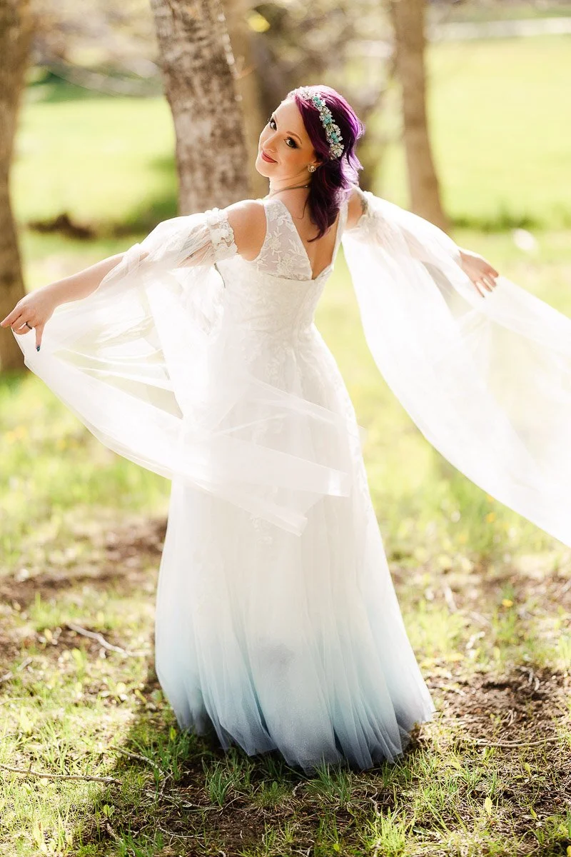 A person in a flowing white dress with flowing sleeves and purple hair poses gracefully in a sunlit forest, conveying a whimsical and serene mood.