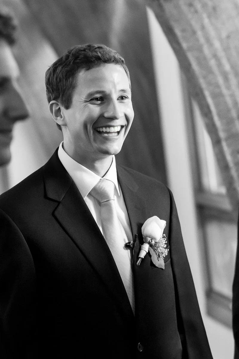 A smiling man in a suit and tie stands indoors. He has a boutonniere on his lapel, exuding joy and excitement, possibly at a formal event. The background is softly blurred.