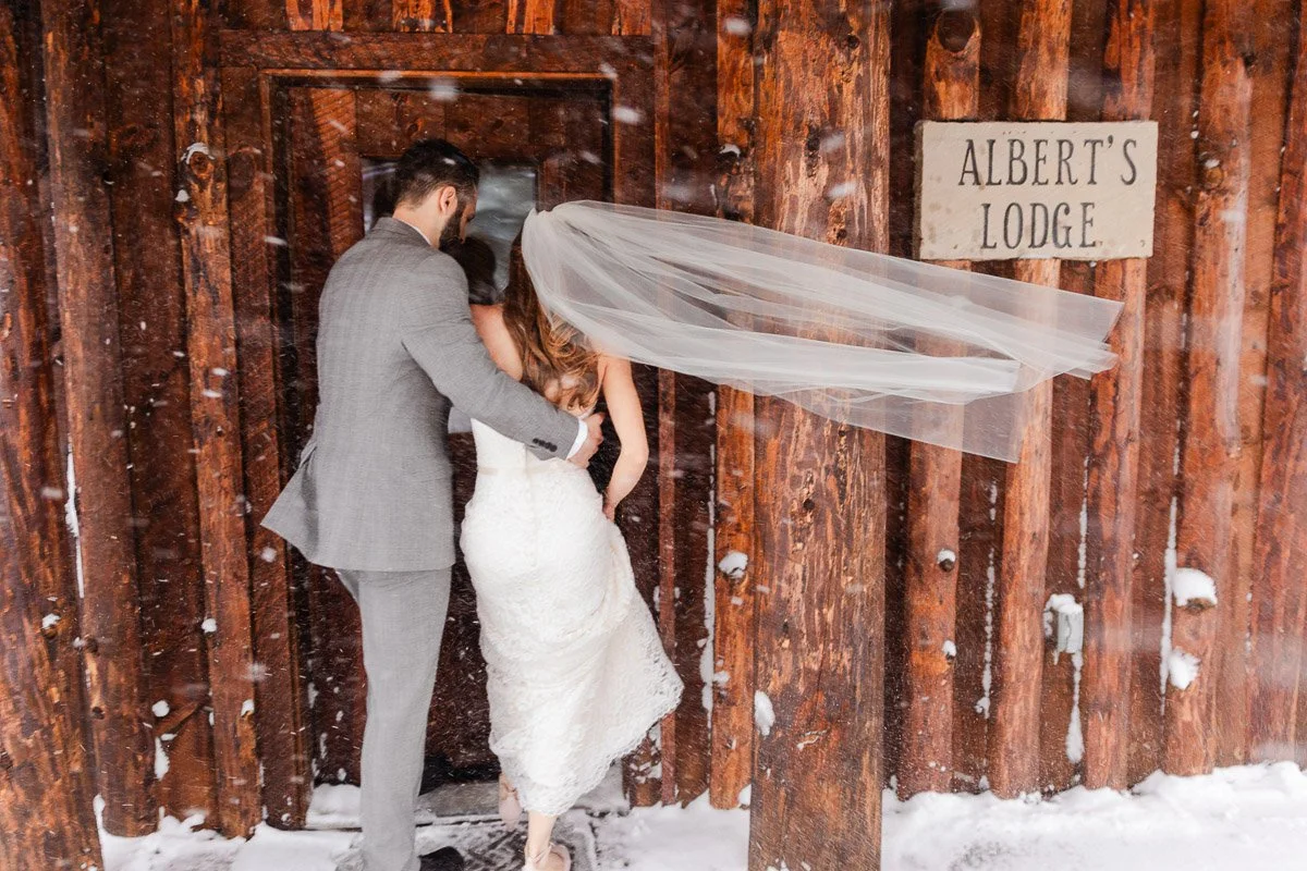 A bride and groom, with the bride's veil blowing in the snowy wind, enter a rustic wooden lodge marked "Albert's Lodge." Cozy, wintery atmosphere during a Spruce Mountain Ranch wedding in Larkspur, Colorado