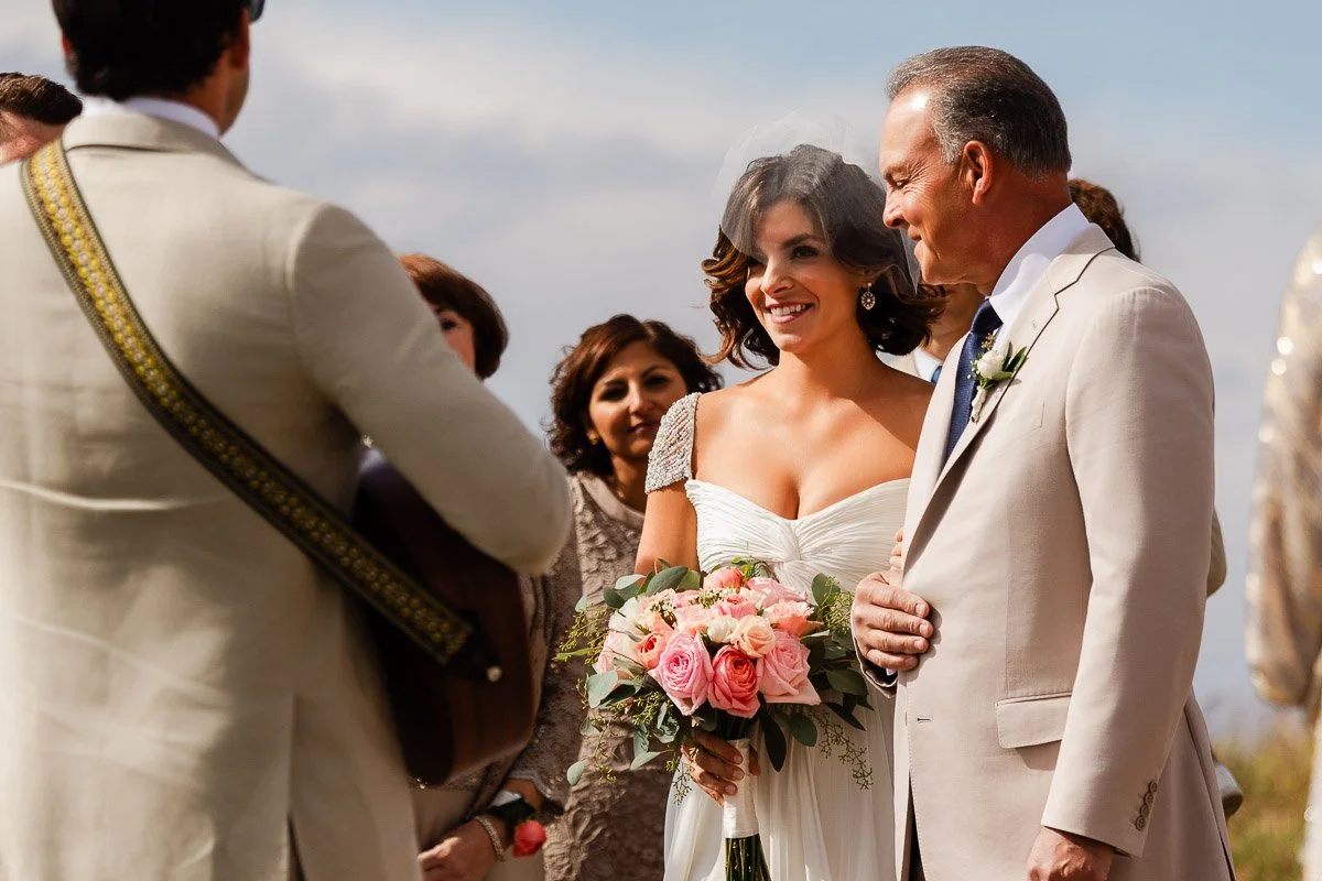 Bride with bouquet and father in light suits, smiling at musician during outdoor wedding. Background shows guests and blue sky, creating a joyful atmosphere.