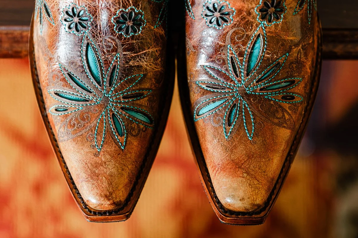 Close-up of two brown leather cowboy boots with intricate teal floral embroidery on the toes, creating a rustic and artistic design.