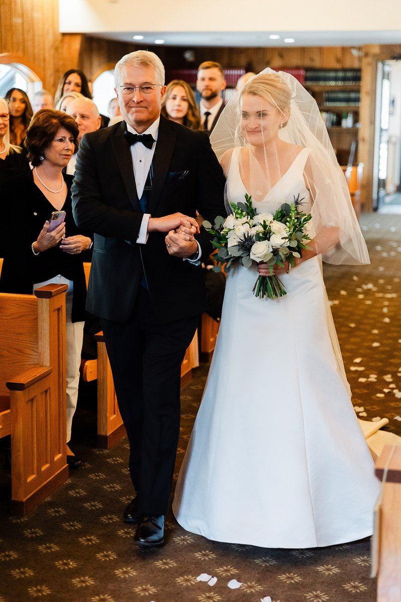 A bride with a veil walks down a church aisle with an older man in a tuxedo, holding a bouquet of white and green flowers. Guests watch with warm smiles.