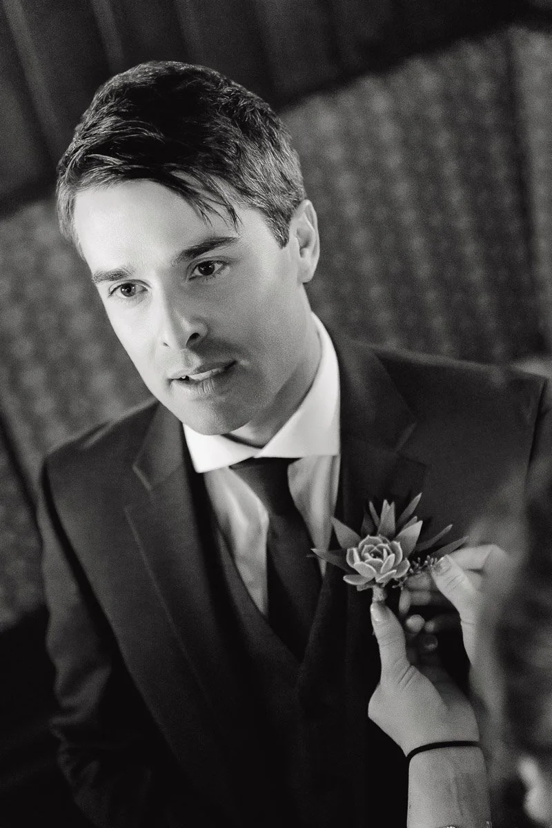 Young man in a suit looking thoughtful, with a hand adjusting a boutonniere on his jacket. The black-and-white image conveys a formal, serene mood.