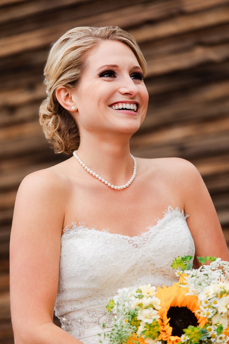 A smiling bride in a lace strapless gown holds a bouquet with a sunflower, set against a rustic wooden background, evoking joy and elegance.