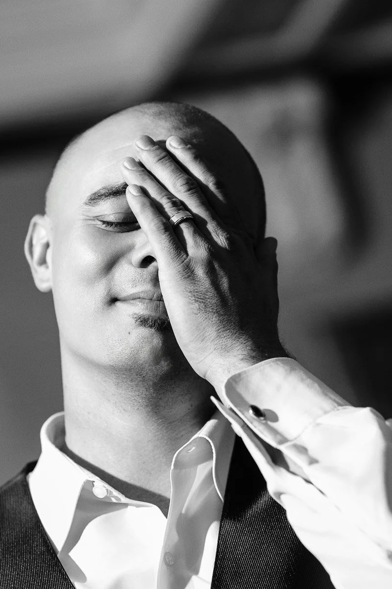 Bald man in a suit, smiling with eyes closed, gently covers face with one hand. The black-and-white setting conveys a serene, reflective mood.