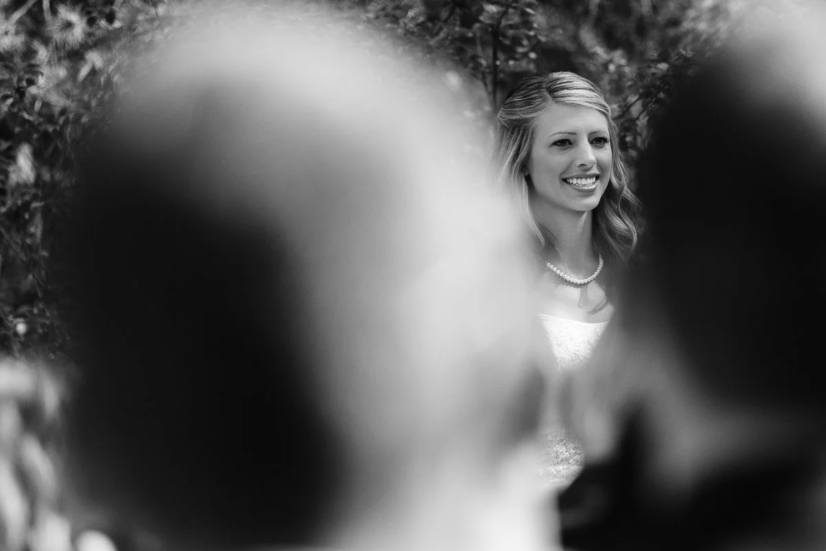 A smiling bride with wavy hair and a pearl necklace stands serenely, framed by blurred onlookers, conveying joy and focus on her special day.