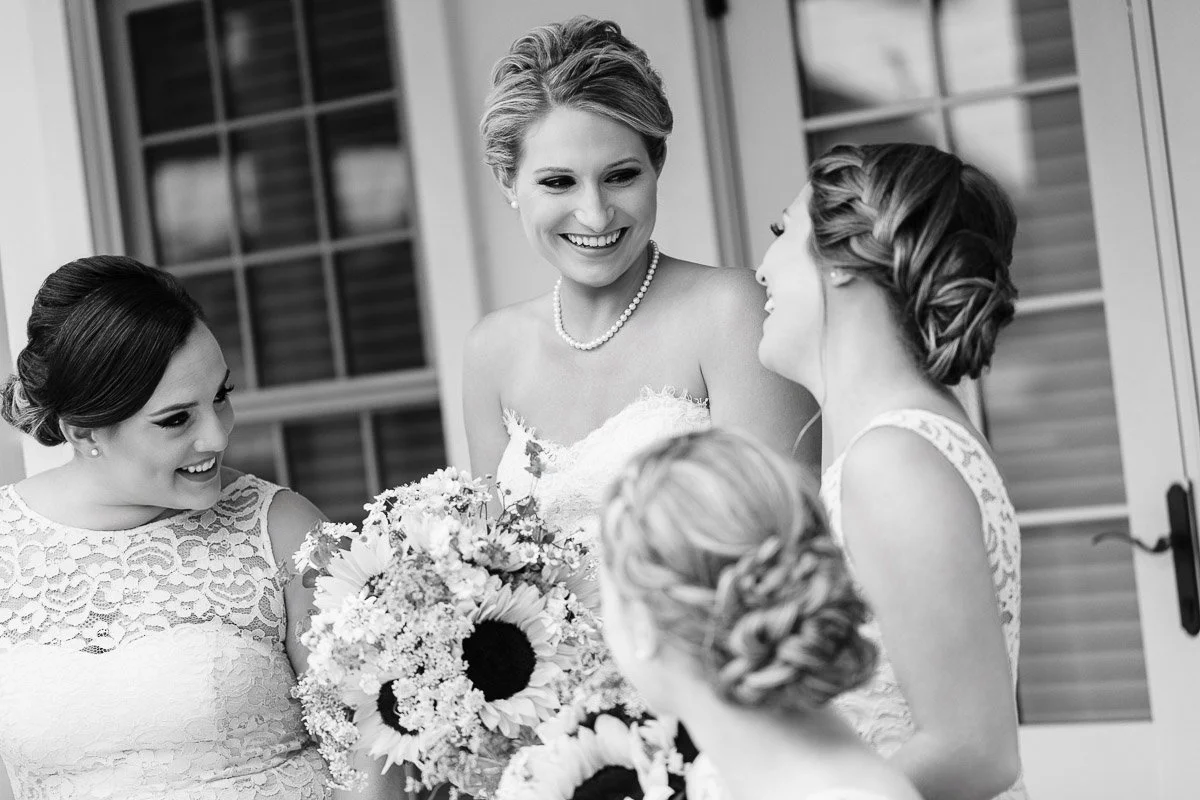 Black-and-white photo of a smiling bride with braided hair, holding a sunflower bouquet, surrounded by joyful bridesmaids in lace dresses.