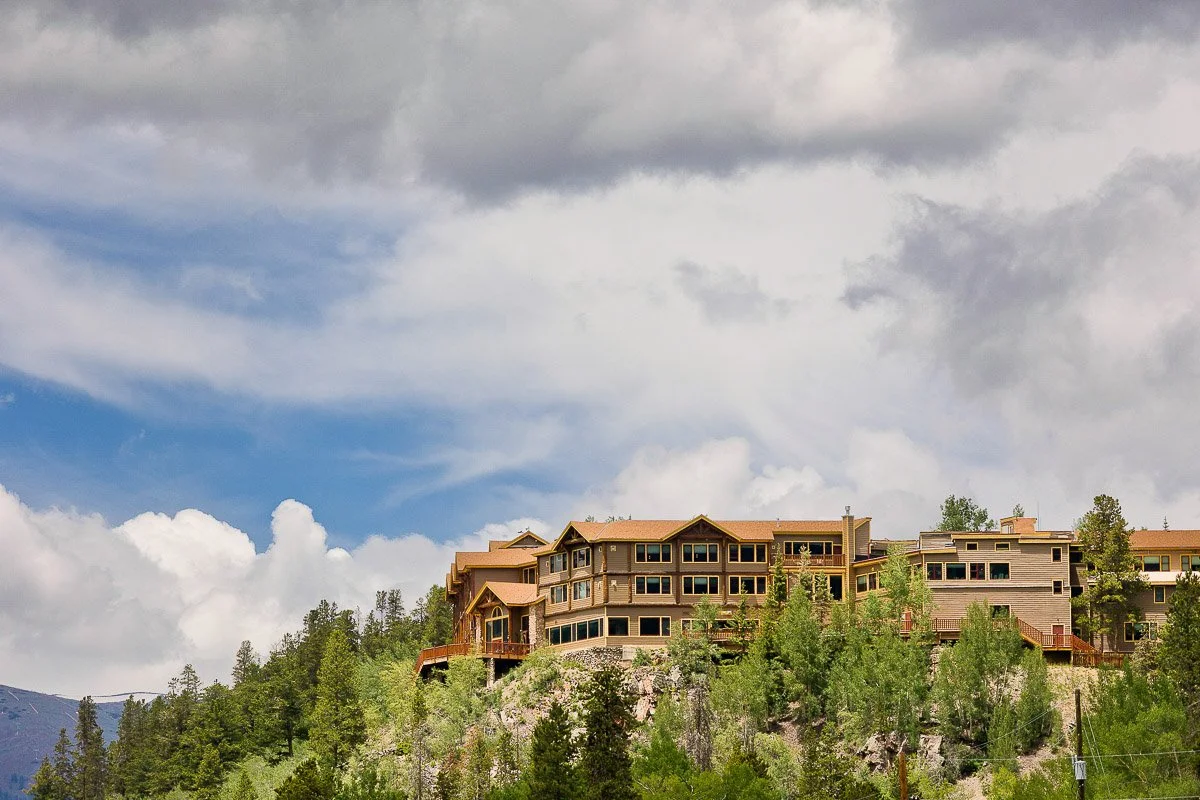 Large mountain lodge atop a forested hill under a cloudy sky, exuding a tranquil, rustic atmosphere. The building is surrounded by pine trees.