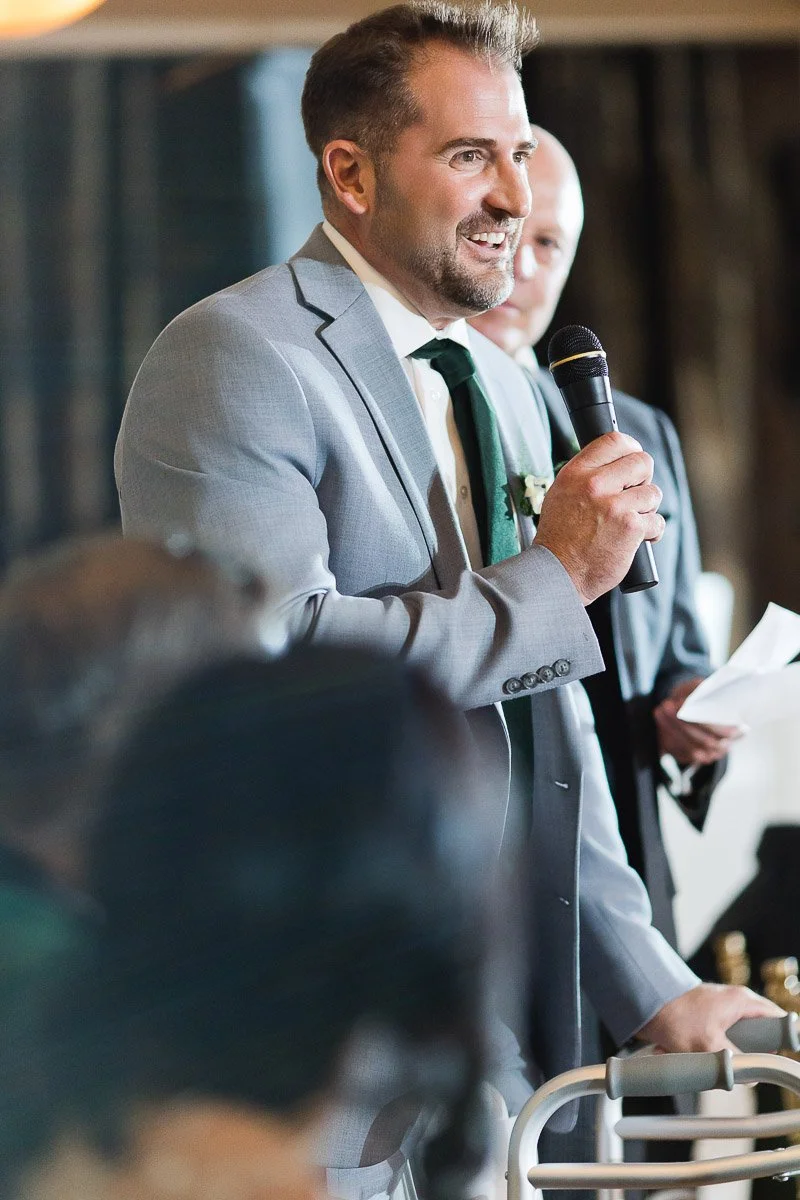A man in a light gray suit and green tie smiles, holding a microphone, speaking at a formal event. The atmosphere is joyful and celebratory.