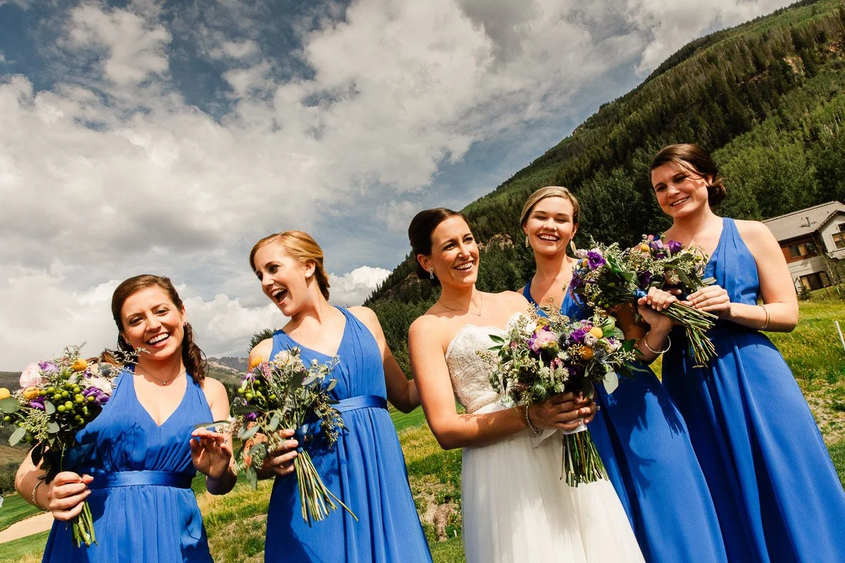 A joyful bride and bridesmaids in blue dresses hold bouquets, laughing under a partly cloudy sky, with a scenic mountain backdrop. Wedding celebration.