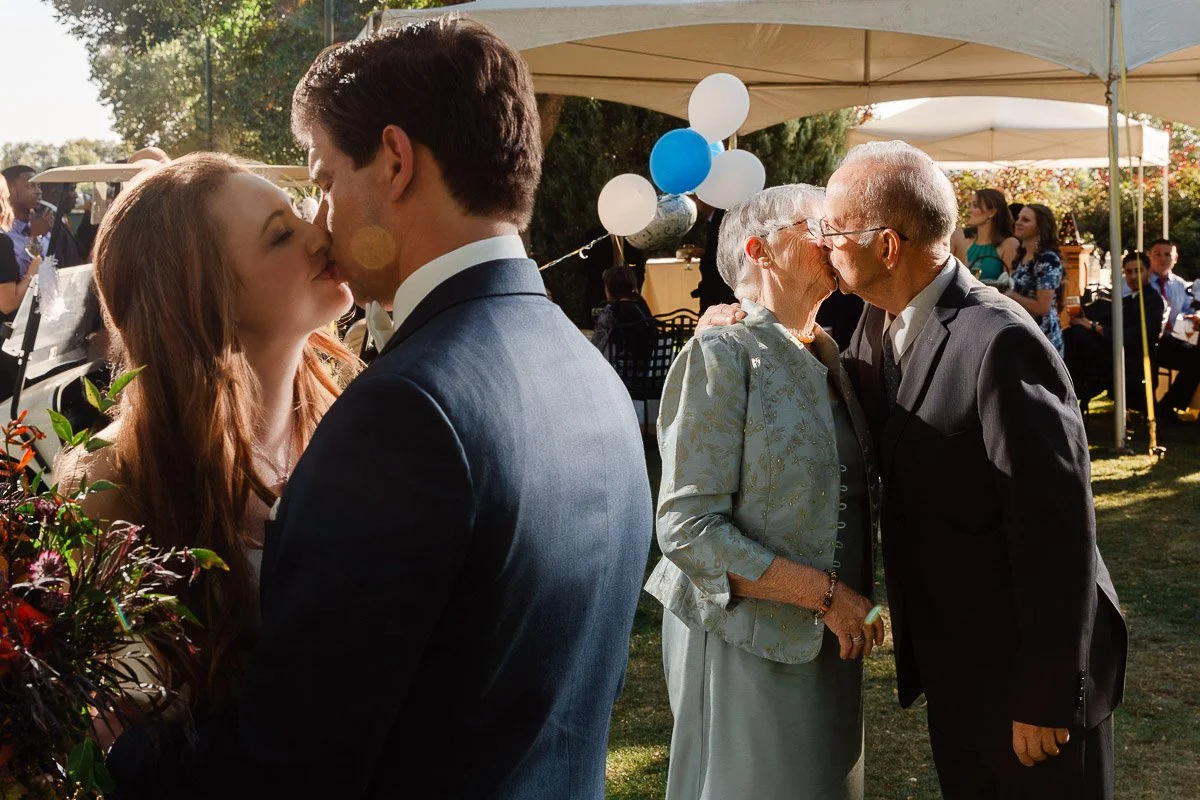 A young couple and an elderly couple share kisses at an outdoor celebration. They are surrounded by trees, balloons, and guests, creating a joyful atmosphere captured by Fort Collins wedding photographer tomKphoto