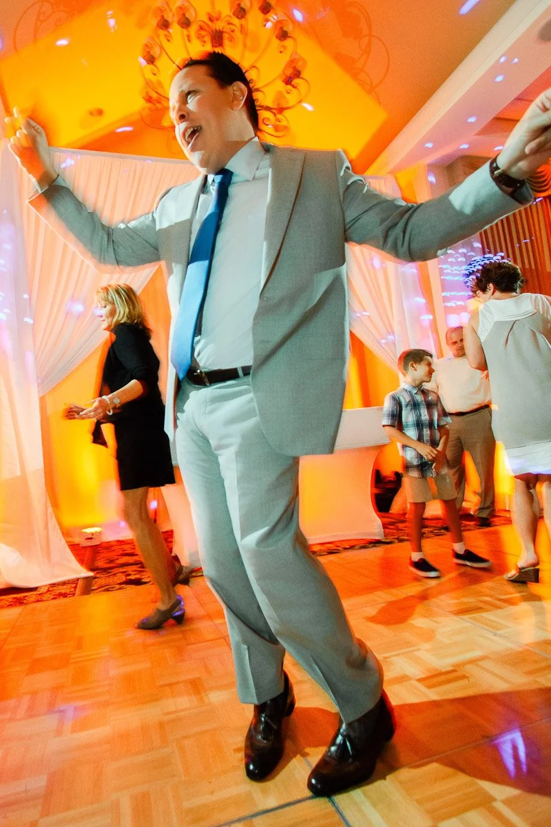 Man in a light gray suit joyfully dancing at a vibrant party with orange and blue lights; guests and a child enjoy the lively atmosphere.