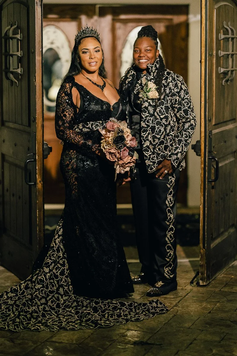 A couple stands smiling in a doorway, adorned in elegant, black, intricately patterned outfits. The woman holds a bouquet and wears a tiara, conveying a regal and joyful atmosphere.