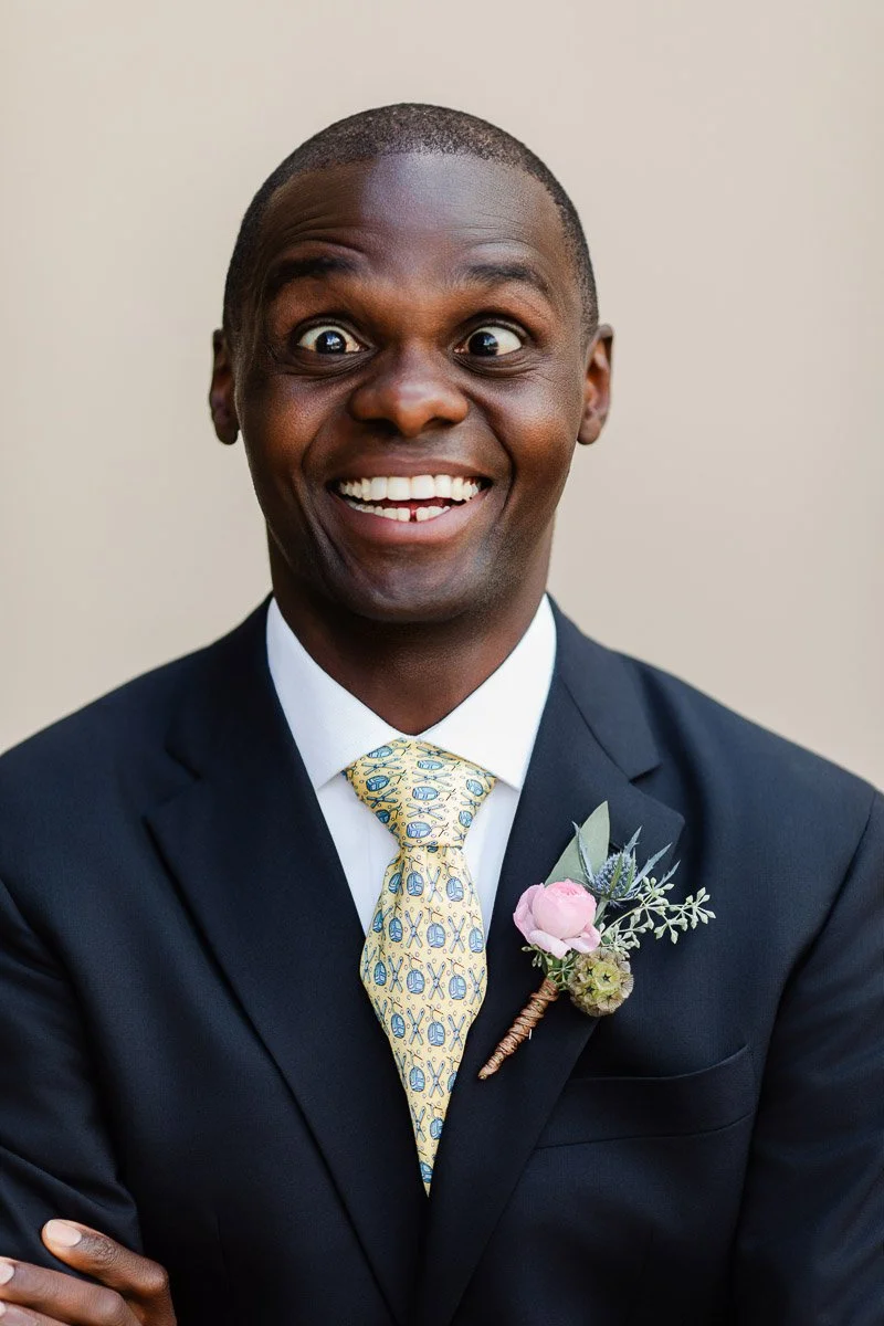 Smiling groom in a navy suit with a patterned yellow tie and floral boutonniere. His wide eyes and expression convey excitement and joy.