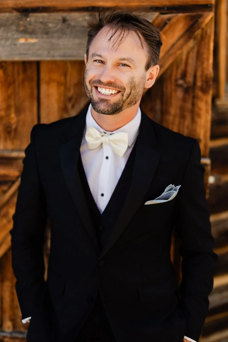 A groom in a black tuxedo and white bow tie smiles warmly, leaning against a rustic wooden wall. The setting is sunlit, adding a cheerful tone during a Strawberry Creek Ranch wedding in Granby, Colorado.