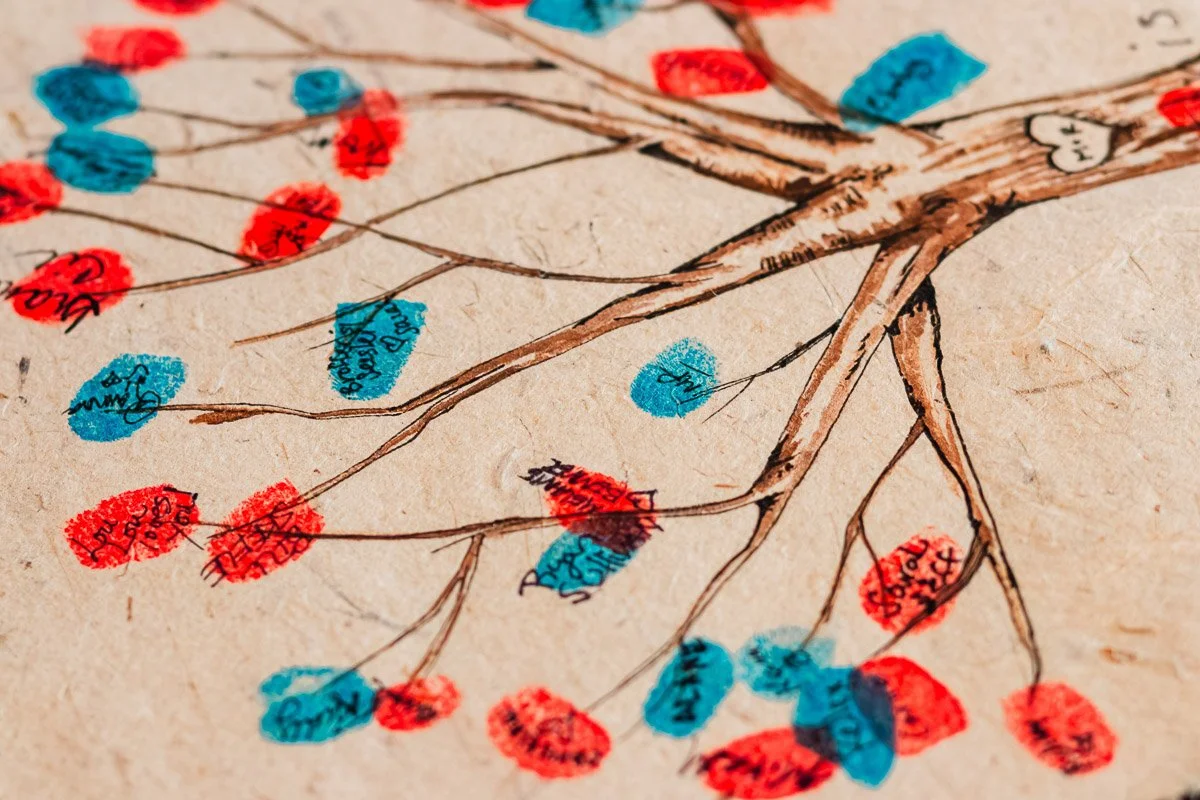 An artistic family tree on paper features branches with red and blue fingerprint leaves. Names are handwritten on the prints, creating a personal touch.