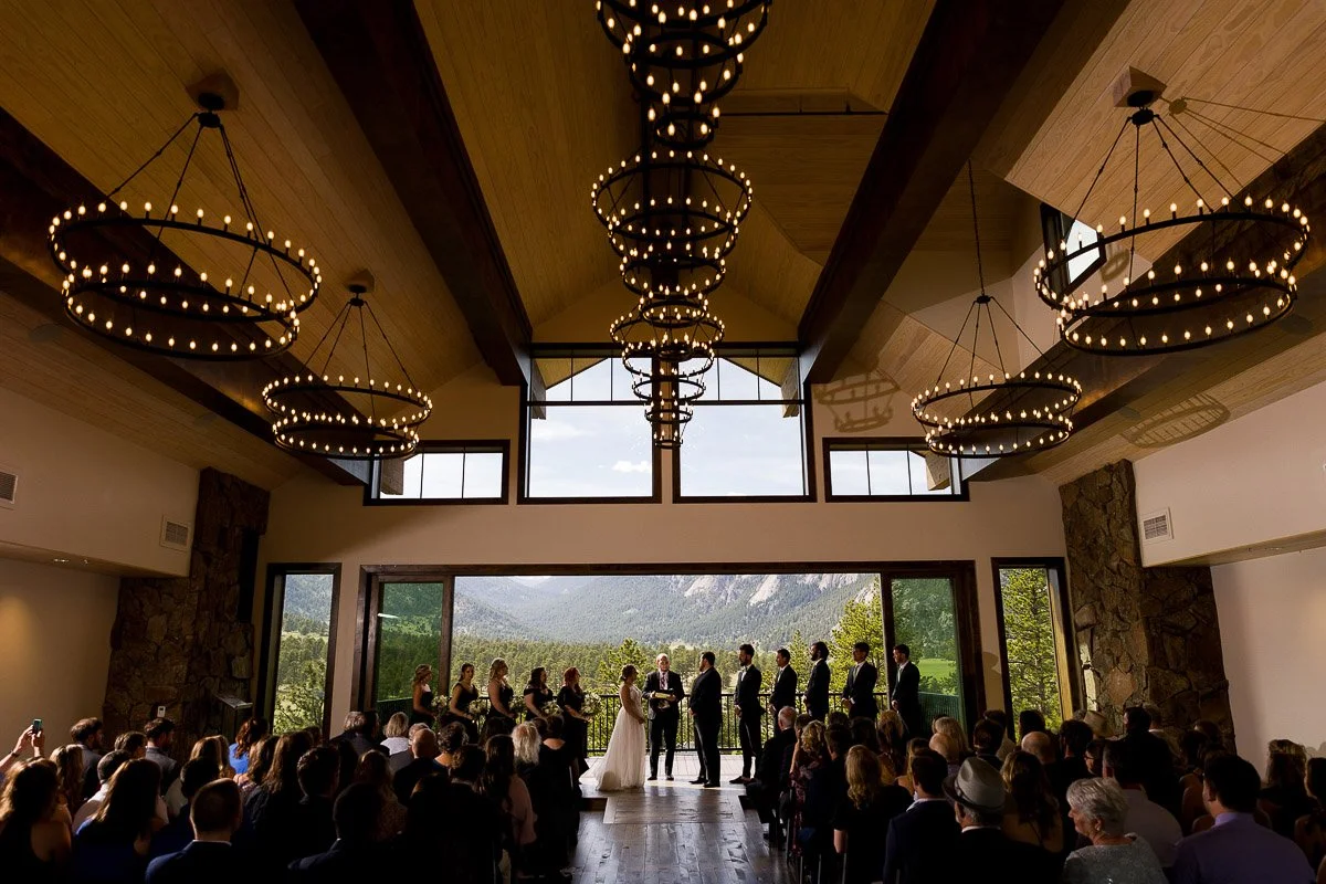 Indoor Black Canyon Inn wedding ceremony with a large window view of mountains, wooden beams, and circular chandeliers. Bride and groom stand at an altar, surrounded by guests.