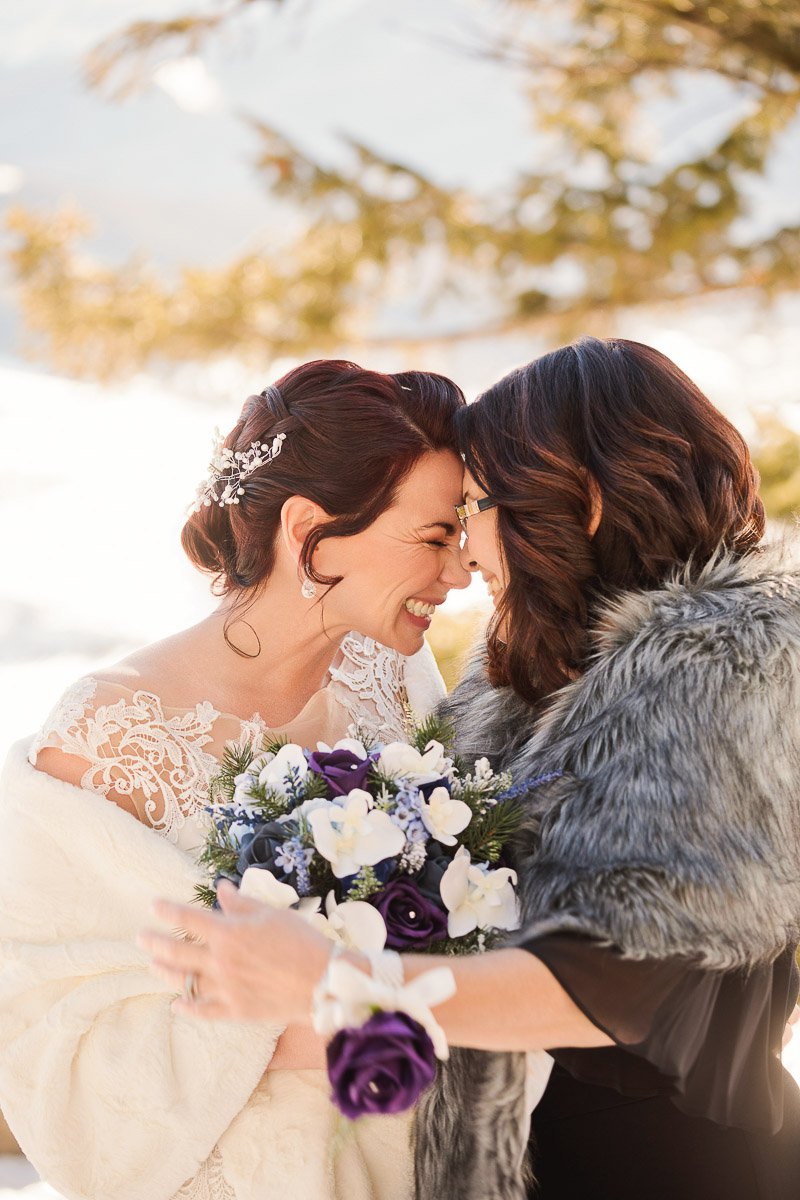 Two women share a joyful moment, leaning in forehead to forehead. One in a lace dress holds a bouquet. The scene is warm and festive.