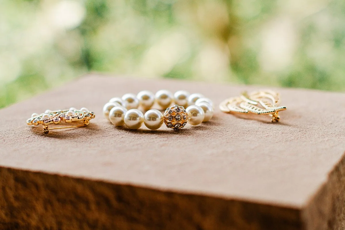 Pearl bracelet and two gold brooches on a textured brown surface, with a blurred green background, conveying elegance and simplicity.
