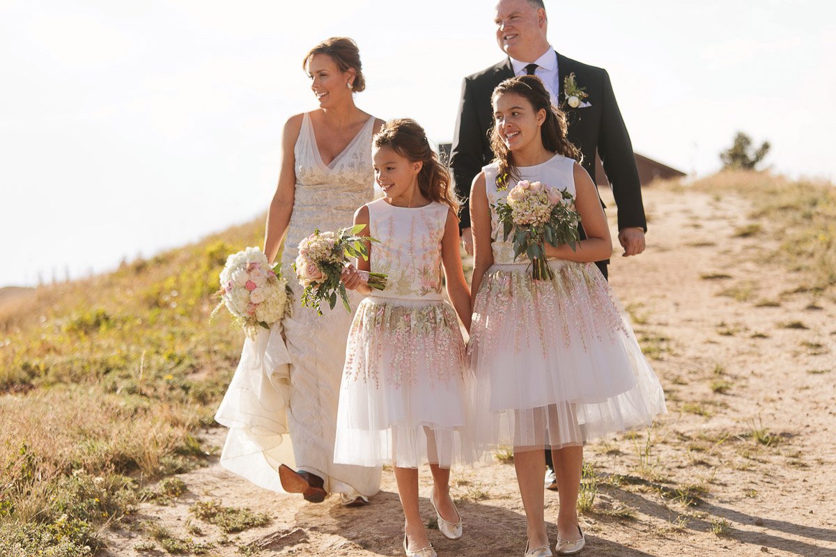 A groom and bride in wedding attire walk on a sunlit dirt path, flanked by two young girls in white dresses holding bouquets, exuding joy and elegance.