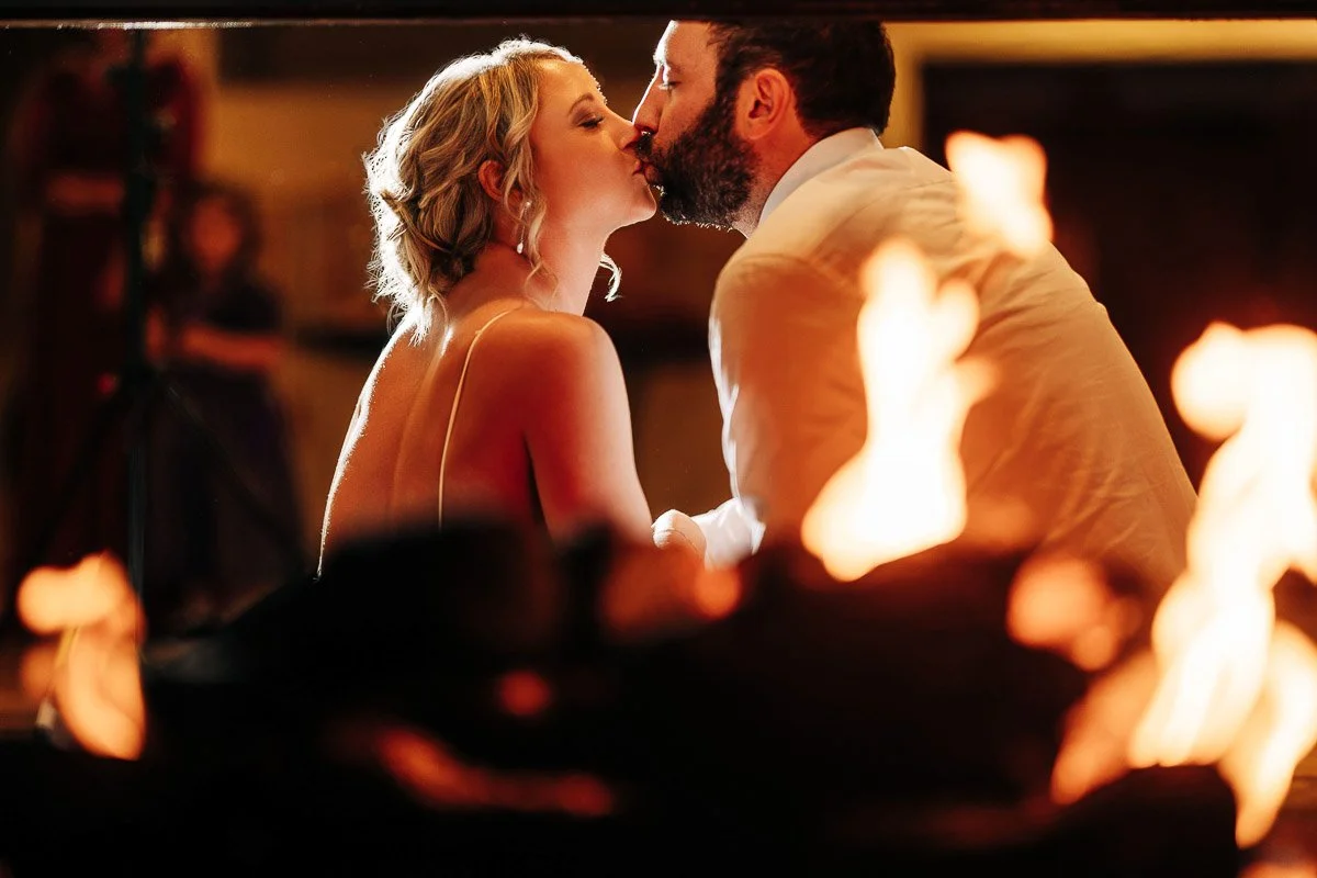 A couple shares a kiss near a softly focused fire, creating a warm and romantic atmosphere. The image exudes love and intimacy.