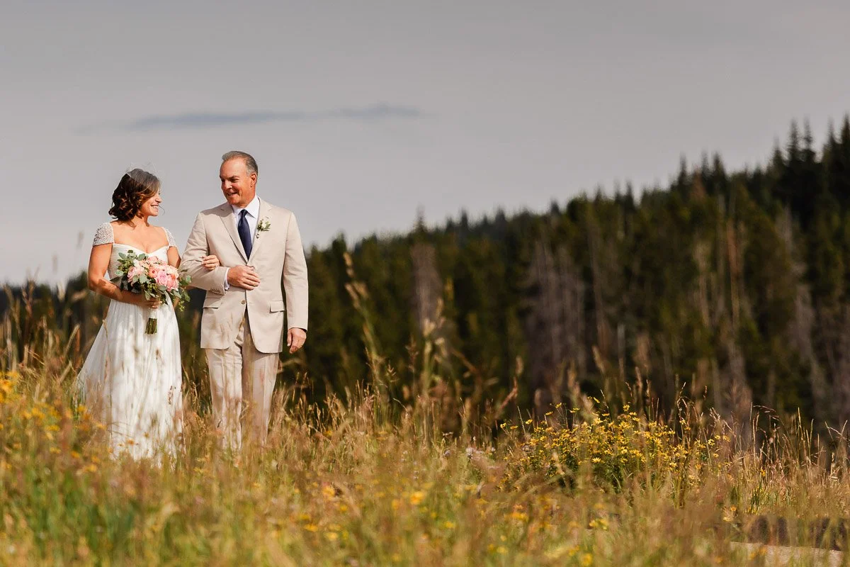 A bride in a white dress holds a bouquet, walking arm-in-arm with a man in a beige suit. They smile amidst tall grass and wildflowers, a forest backdrop.