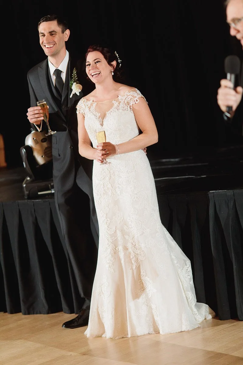 A joyful bride and groom stand together on a stage, holding champagne glasses. The bride wears a lace wedding dress; the groom is in a black suit.