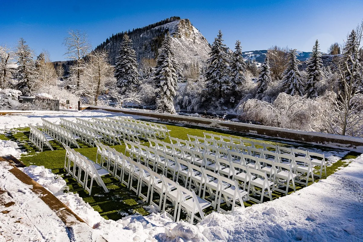 Rows of white chairs on green grass are set for an outdoor Westin Riverfront Resort wedding, surrounded by snow-covered trees and mountains under a clear blue sky.