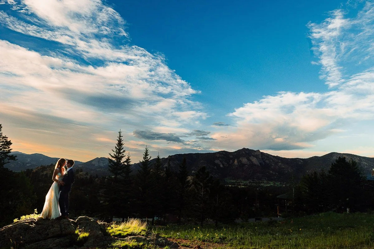 A couple embraces on a rocky hilltop at sunset, surrounded by pine trees and overlooking distant mountains under a vibrant blue sky with wispy clouds.