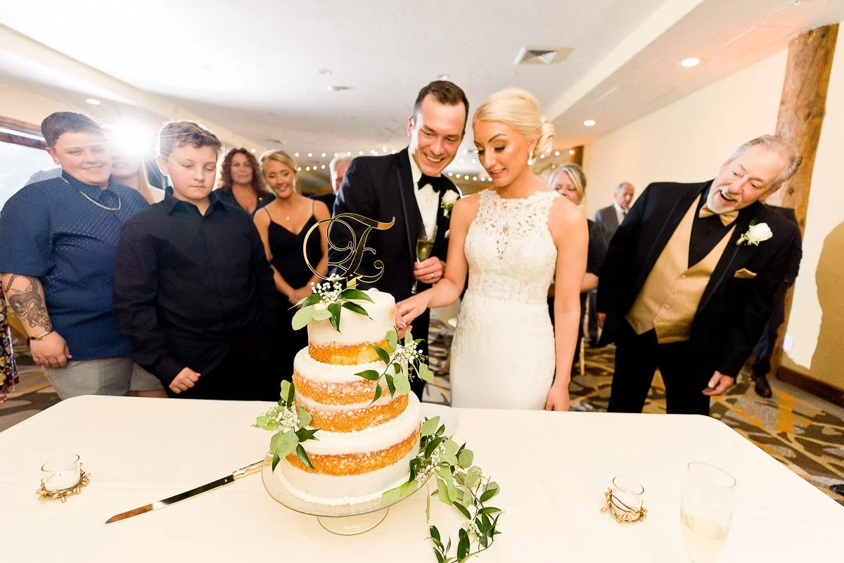 A joyful bride and groom cut a tiered semi-naked wedding cake adorned with greenery. Guests watch with smiles and excitement in the warmly lit room.