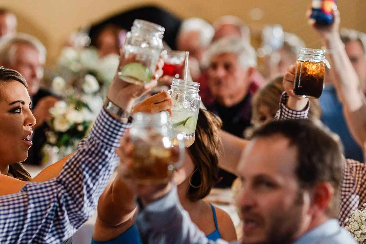 A group of people at a festive gathering raise mason jars with various drinks in a cheerful toast, surrounded by a warm, lively atmosphere.