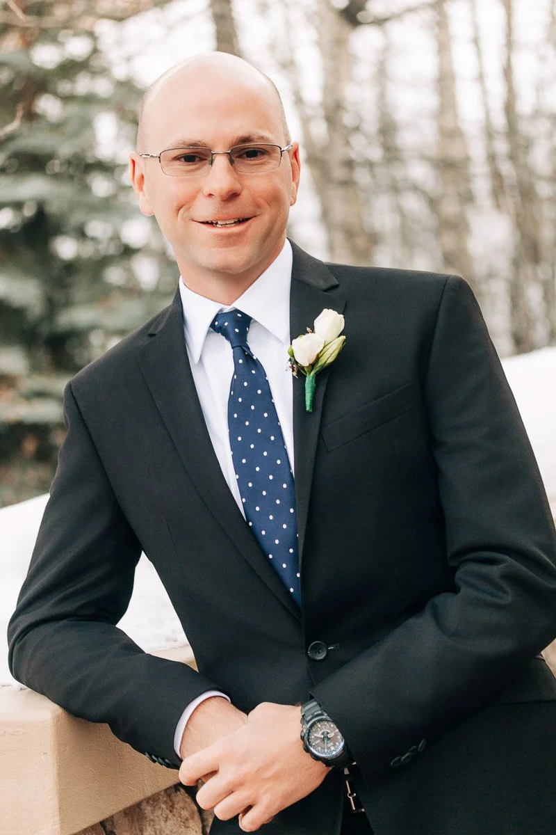 A man in a black suit and blue polka dot tie leans on a ledge, wearing glasses and a boutonniere. He smiles warmly, with snowy trees in the background.