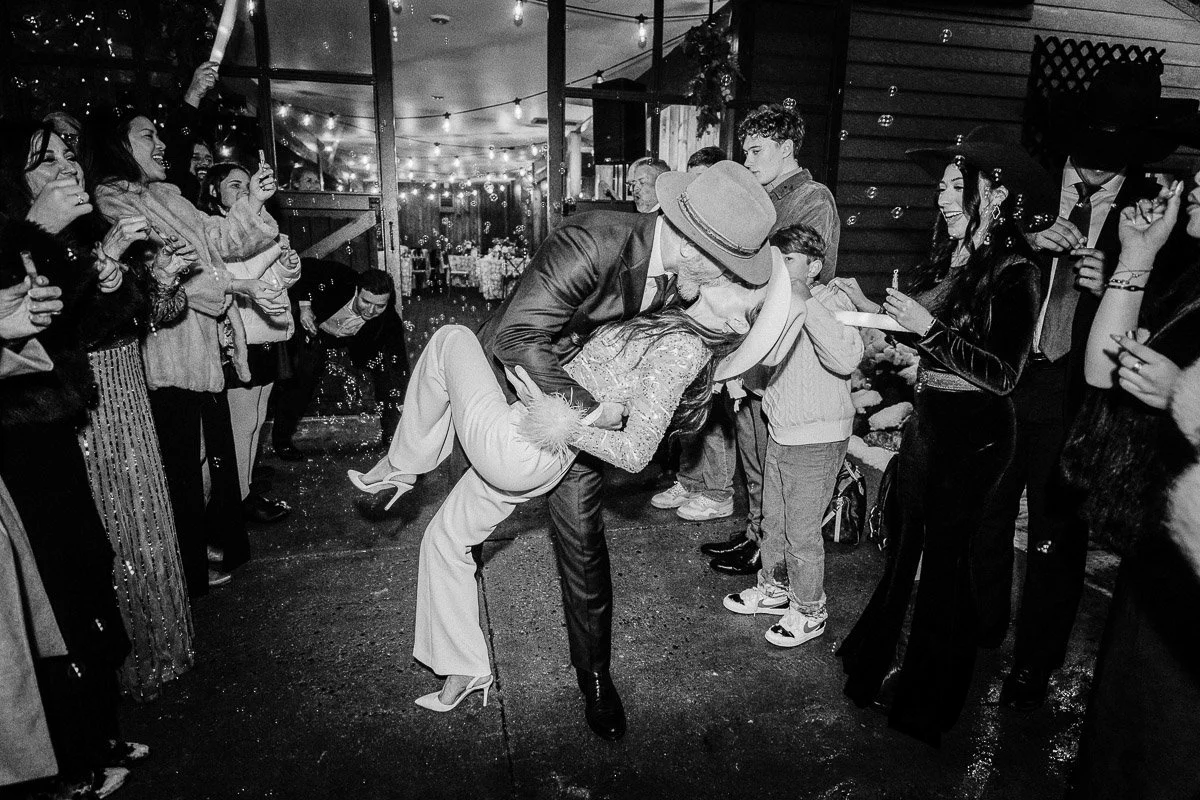 A black and white photo shows a man in a suit and hat dipping a woman in glittery attire for a kiss. They are surrounded by a joyful crowd under string lights, exuding celebration and romance.