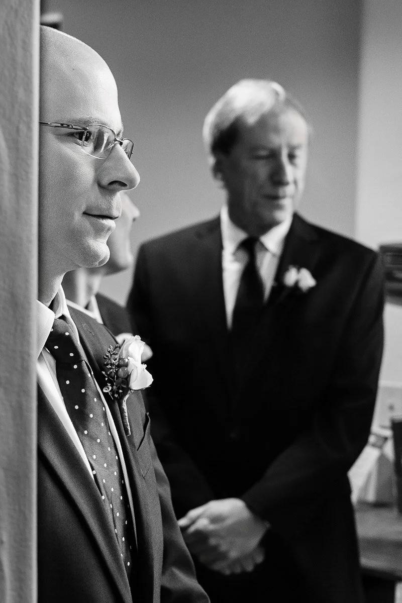 Black and white photo of two men in suits, one in focus with glasses, a polka dot tie and boutonniere, and the other blurred in the background, both appearing thoughtful.