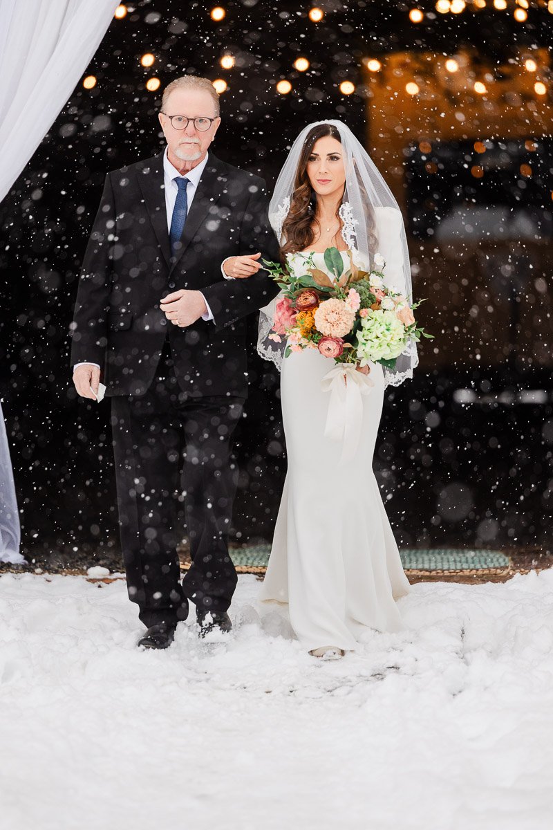 Bride in white gown and veil walking with a man in a black suit through falling snow, holding a colorful bouquet. Warm, festive lights glow softly in the background.