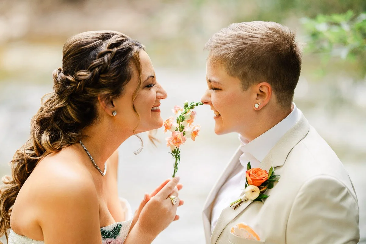 A couple in wedding attire shares a joyful moment. The person on the left has long hair and a white dress, and the person on the right, in a white suit, holds pink flowers.