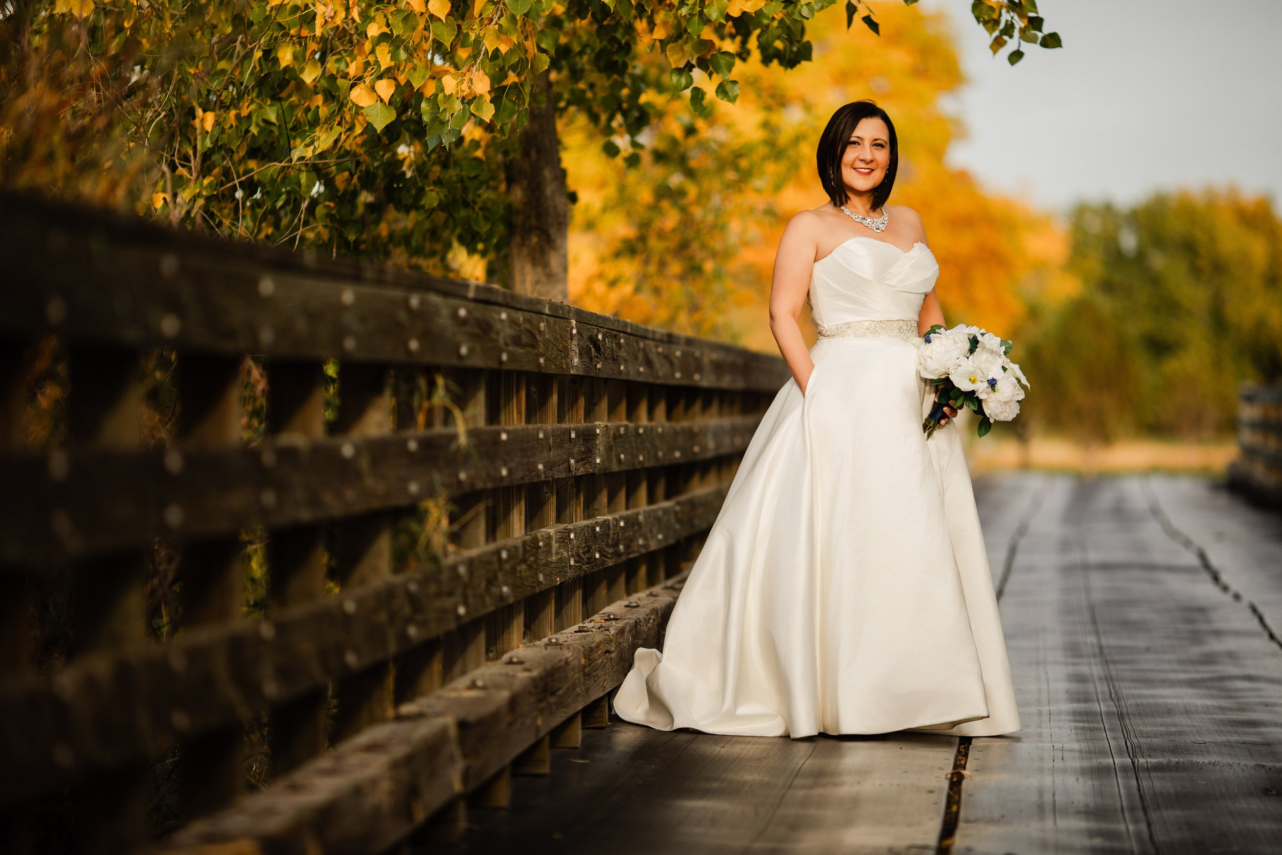 Bride in a white gown stands on a wooden path lined with autumn trees, holding a bouquet. The scene conveys a serene and joyful atmosphere during a Valley Country Club wedding in Aurora, Colorado.