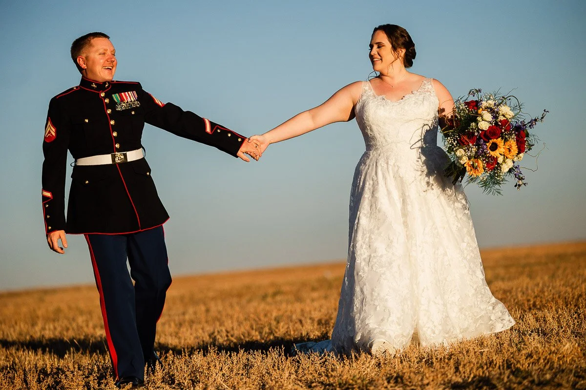 A bride in a white lace gown holds hands with a groom in a military dress uniform. They smile at each other in a sunlit field, conveying joy.