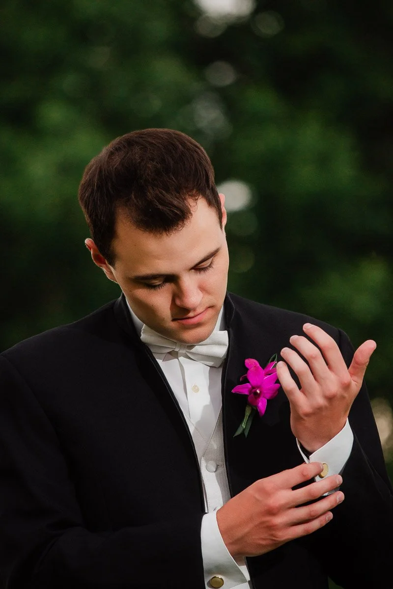 Young man in a black tuxedo adjusts his cufflink, wearing a white bow tie and purple boutonniere. Background is blurred greenery, creating a formal, serene mood.