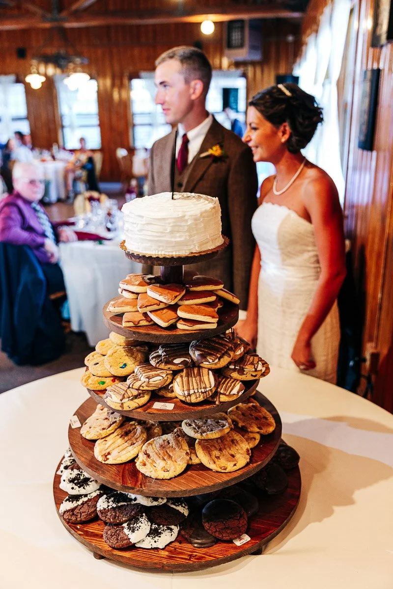 A bride and groom stand beside a tiered dessert display featuring a variety of cookies and a small cake on top. The setting is a cozy wedding reception.