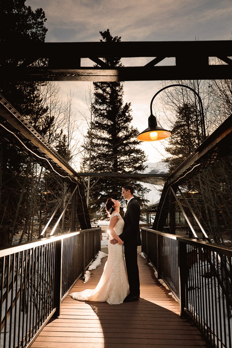 Bride and groom stand on a snowy bridge at sunset, sharing an intimate embrace. Warm glow from a lamp casts a romantic light, with trees silhouetted during a Silverthorne Pavilion Wedding.