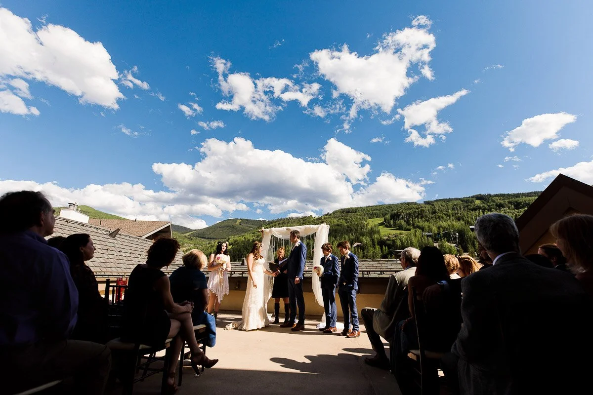 A couple exchanges vows under a floral arch on a sunny terrace with mountains in the background. Guests watch, creating a joyous, serene atmosphere.