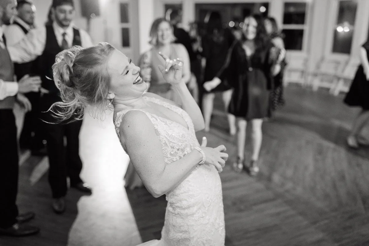 A joyful woman in a lace dress laughs while dancing at a party, surrounded by people in formal attire. The scene exudes happiness and celebration.