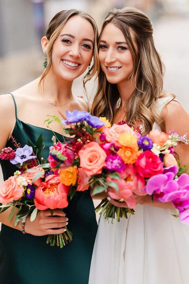 Two smiling women hold vibrant bouquets of colorful flowers, including roses and orchids. One wears a green dress, the other a white dress, conveying joy.