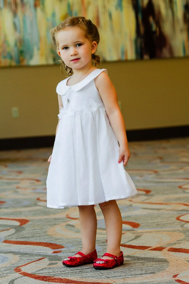 A young girl wearing a white dress and red shoes stands on a patterned carpet. Her hair is styled with curls, and she appears calm and curious.