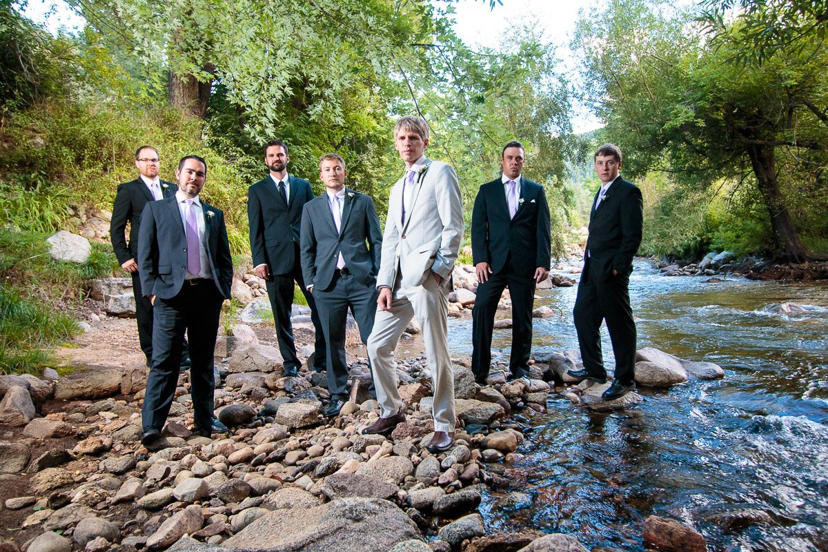 A group of men in suits stand confidently on rocky terrain beside Boulder Creek, surrounded by lush green trees, conveying a formal yet relaxed vibe.