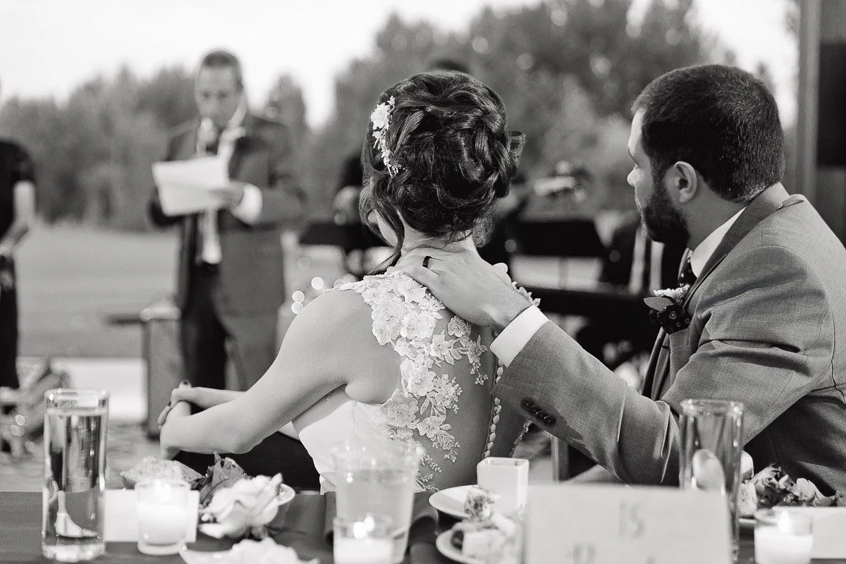 A couple, seated at a table, listens intently to a speech at a wedding. The groom gently holds the bride's shoulder. The setting is elegant and intimate.