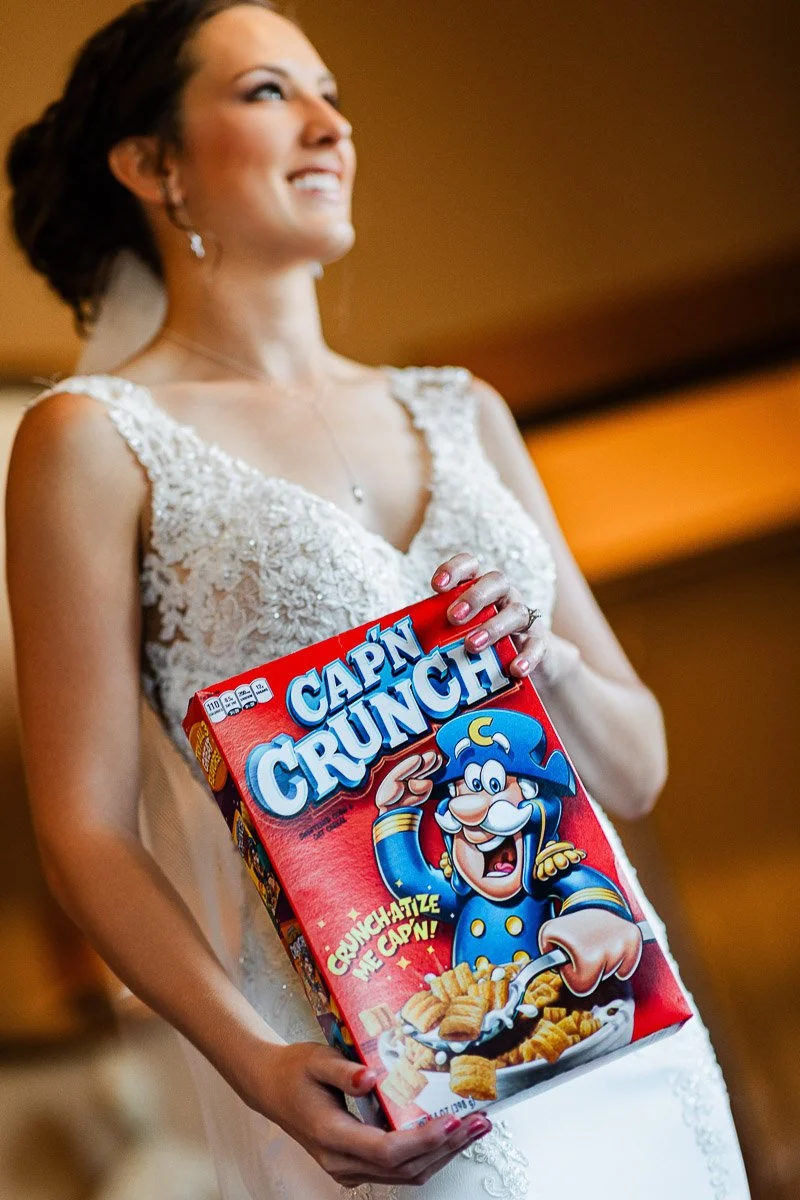 Bride in white lace dress joyfully holds a Cap'n Crunch cereal box, featuring the cartoon character, creating a playful and lighthearted moment.